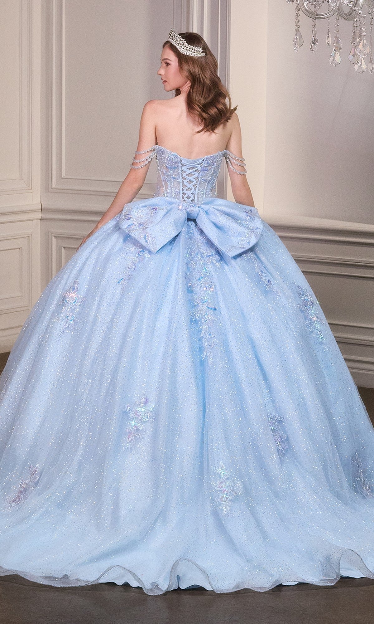 A person stands in an elegant room wearing the Quinceanera Dress 15735 By Ladivine, a sparkling light blue ball gown with an off-the-shoulder neckline, a large bow on the back, and a tiara.